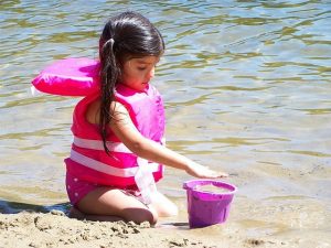 Young girl in pink life vest playing in sand at beach near water