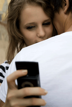 Boy is hugging girl as she looks at the cell phone she is holding behind his shoulder.