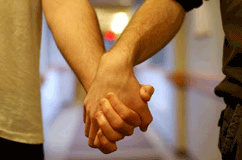 Here is a partial side view of two people wearing T-shirts and pants holding hands, with the focus on their clasped hands.