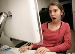 Girl is sitting in front of computer monitor with shocked look and mouth open wide.