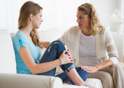 Mother and daughter are sitting on couch; mother touches daughters knee as she looks at her and talks.