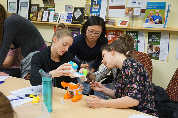 Three individuals sit at a table. They are all huddled around a children's toy which is opened up.