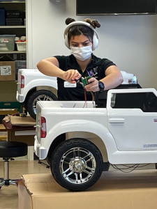 A person wearing a white mask works on the wiring in a white ride-on car during a workshop