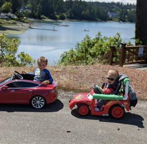 Two children in red ride-on cars in front of a lake. Both children are looking at the camera and one is wearing sunglasses.