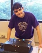 Photo of Alexander. He is wearing a purple shirt and standing over top of an adapted xbox controller that he made while on a design team.