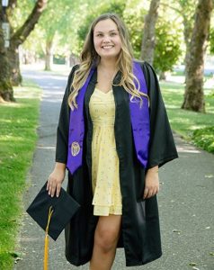 Photo of Anyssa. She is wearing a graduation robe and holding a graduation cap standing outside on a path lined with trees.