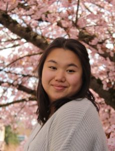 Photo of Jaelynn. She has shoulder length black hair and is wearing a white shirt. She is standing in front of a blooming cherry blossom tree.