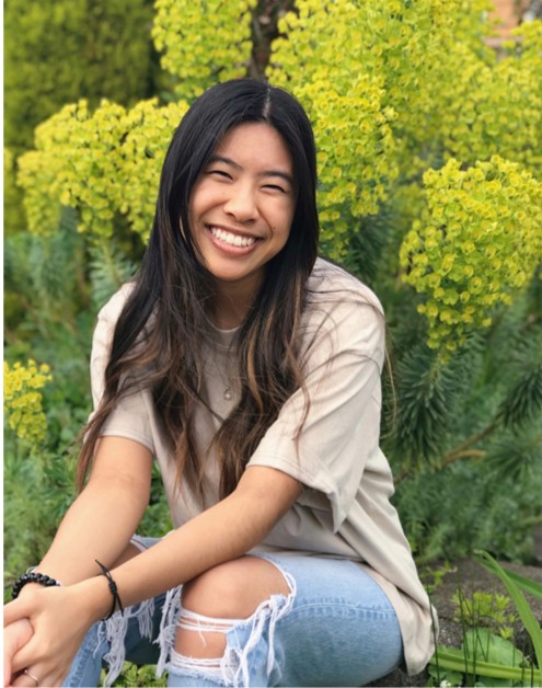 Kaitlin Danh Photo of Kaitlin. She is wearing ripped jeans and a white shirt and sitting in front of green plants.