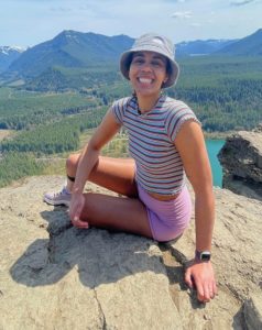 Kiana Keithley Photo of Kiana. She is wearing a bucket hat, stripped shirt and pink shorts and sitting on a rock with mountains in the background.