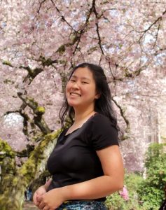 Maelynn Danke Photo of Maeylnn. She is wearing a black shirt and standing in front of a tree with white cherry blossoms.