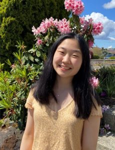 Maia Song Photo of Maia. She has shoulder length black hair and is wearing a yellow shirt and standing in front of bright pink flowers.