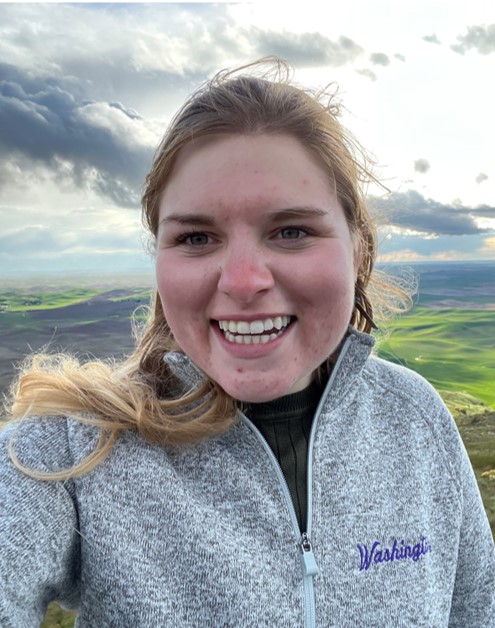 Photo of Mia. She is wearing a grey pull over sweatshirt and has blond hair pulled back. She is standing in front of a vista of rolling green hills.