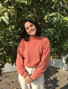 Photo of Mishti. She has shoulder length black hair and is wearing a salmon colored shirt and standing in front of a tree.