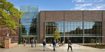 Brick multistoried university building with large windows on the front and a large windowed atrium entrance. Students walk into the building through the main entrance.