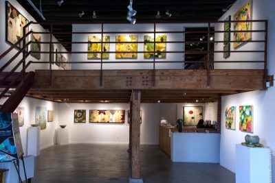 White walled gallery space, with a lofted second floor space. The walls are filled with paintings, and there are a few 3 dimensional pieces on pedestals.
