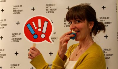 Person with light skin wearing a yellow cardigan stands in front of a white backdrop with a tiled black lettered logo reading "School of Art + Art History + Design." They are eating a small cupcake and holding up a red speech bubble sign with exclamation marks.