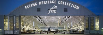 Open airplane hangar at night, brightly lit from the inside and featuring multiple airplanes. Lit text sign above the entrance reads "Flying Heritage Collection"