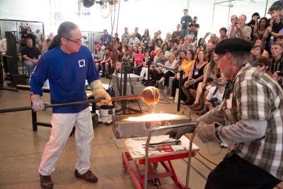 Two people blowing hot glass in front of a crowded audience.