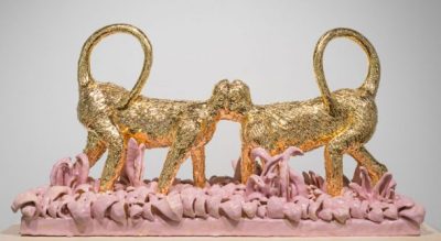 Ceramic statue featuring two gold monkey figures, their faces meeting in the middle and their tails mirrored. The figures stand on a shiny, pink, grass-textured base.