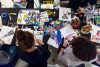 Overhead view of youths working at a crafting table with collage materials. Two central youth figures are cutting things out of magazines.