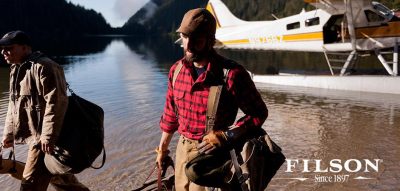 Two ruggedly dressed masc people exit a plane floating on a river, each carrying gear. Text in the lower right corner reads "Filson; Since 1897"