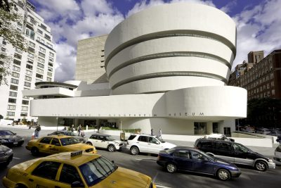 Modern art building from the outside. The building is smooth white stone, with a circular funnel shaped multi-story top. Sign out front reads "The Solomon R Guggenheim Museum"