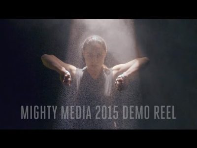 Person with an intense gaze walks through a water fall of sand, their hands raised as though they are parting a curtain. Text across image reads "Mighty Media 2015 Demo Reel"
