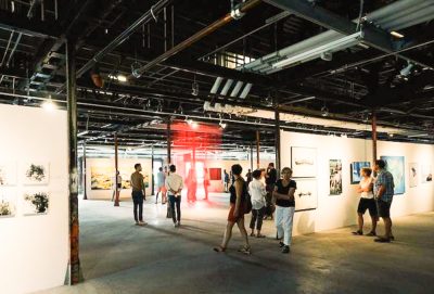 Open art exhibition space with free standing white walls upon which 2D artwork is hung. Small groups of people walk around the space, viewing art.