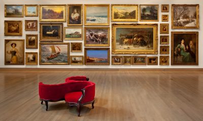 Bright red modern chair in front of a gallery wall with multiple framed paintings displayed salon style.