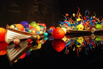 Colorful glass art installation; glass orbs and organic, vine-like forms are arranged inside two canoes on a polished black surface.