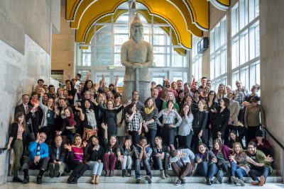 Group photo of museum staff gathered in a brightly lit museum front foyer, around a large multi-story indigenous figure sculpture