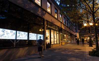 Sidewalk featuring a row of several storefront/gallery front spaces at dusk. Two people walk down the sidewalk, and behind a single person views a video installation in some windows