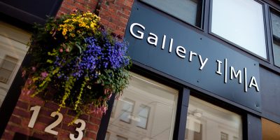 Metal sign over a brick and glass store front reads "Gallery IMA", a basket of bright flowers to the left of the sign.