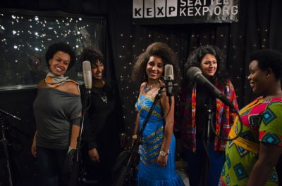 Group of five femme people in a recording studio in front of several microphones, all smiling at each other and singing