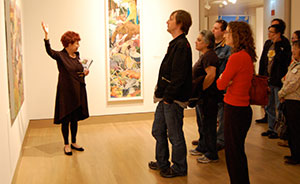Museum docent speaks in front of a group of museum goers, gesturing to an art piece on the wall.