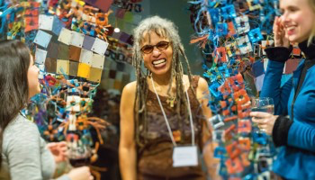 Person with dreadlocks and glasses grins in front of a multi-media art sculpture.