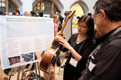 Person showing details of a guitar in front of a presentation board, in a crowded research fair setting.