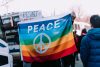 Person holds a rainbow flag with a white peace sign and text reading "Peace" outside in a protest march.