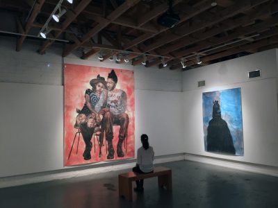 Person sits on a bench in a darkened art gallery, in front of a large painting of two figures