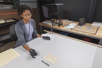 Museum intern wearing gloves points to an archived document on a table with a metal instrument.