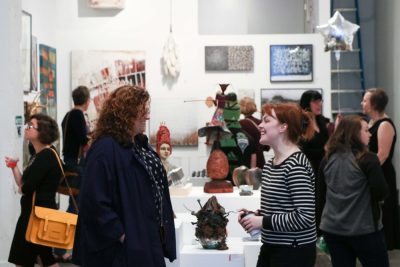 Gallery opening featuring both 2D work hung on the walls and 3D work displayed on pedestals. People mingle and chat in the background, and two femme people stand in the foreground talking and smiling.