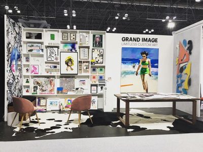 Staged fair booth with a salon-style display wall of different types of artwork. Two chairs sit in front of the wall, and a table to the right holds more 2D artwork. Sign on the left right of the booth reads "Grand Image: Limitless Custom Art"