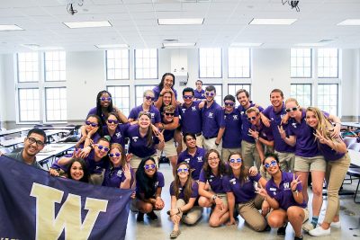 Group of students in matching purple UW polos and khakis, most wearing sunglasses. Two are holding a UW flag in the bottom left corner.
