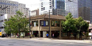 Exterior of a two story office building on a street corner, skyscrapers visible behind.