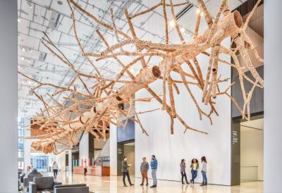 Large organic tree sculpture installed in a museum main foyer. Small groups of people stand below chatting.