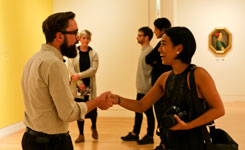 Two smiling people shake hands in an art gallery setting.