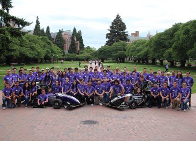Group of students all wearing matching purple organization t-shirts stand and sit in front of the University quad, two formula-1 race cars in front.
