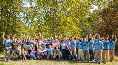 Group of people all wearing the same blue company shirt pose together outside, their hands raised in a celebratory fashion.