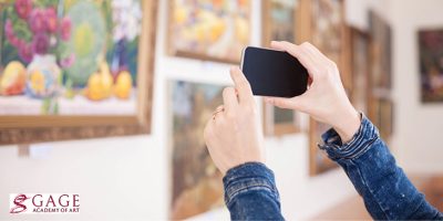 Hands holding a smart phone, taking a picture in an art gallery setting. Text in lower left corner reads "Gage Academy of Art"