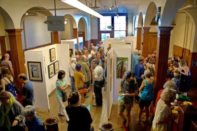 Busy art gallery from above, with many groups of people milling about and looking at artwork on the walls.